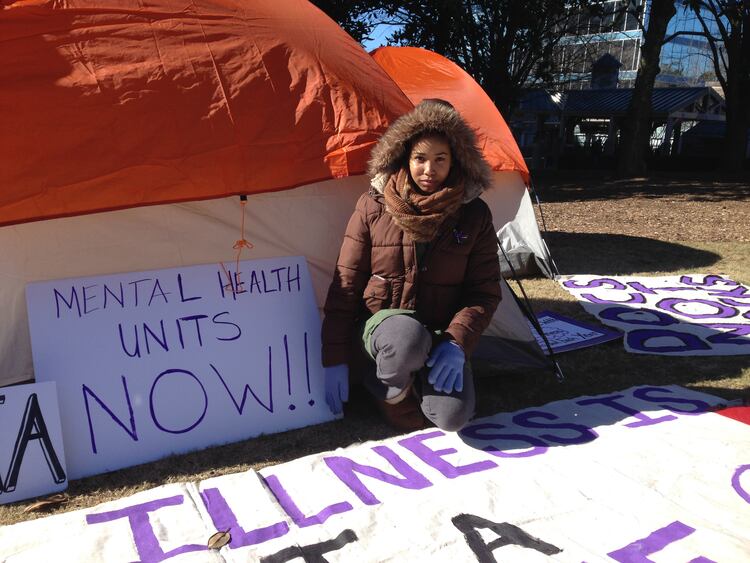 Bridget Anderson kneels in front of a tent with a hand written sign "mental health units now" and she is wearing a thick brown winter coat with a fuzzy hood